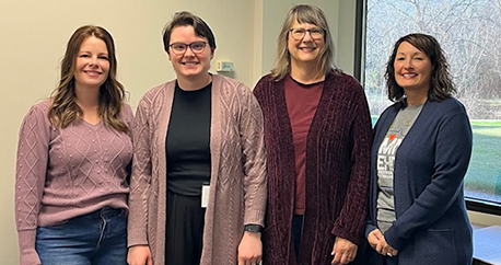 Four white women are standing together and smiling. They are wearing sweaters