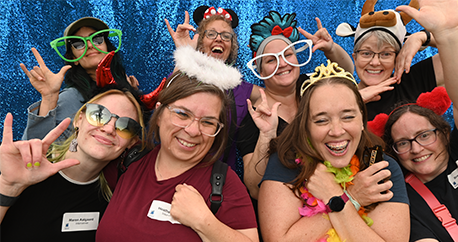 A group of 8 women who are all community interveners are making fun faces and wearing photo booth props