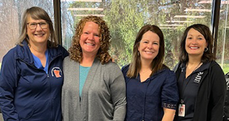 Picture of four white women standing in front of a glass window smiling at the camera.
