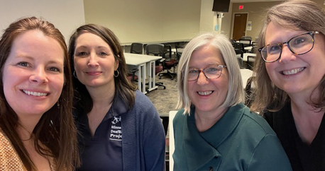 Picture of four white women standing together in a training room smiling at the camera.