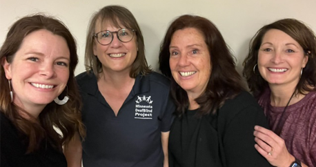 From left to right are Brandy Sebera, Ann Mayes, Carolyn Monaco, and Deanna Rothbauer. All four are white women standing closely together in front of a white wall and smiling at the camera.