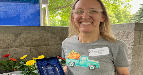 A white, middle aged woman with blonde hair and glasses is standing outside with flowers and smiling
