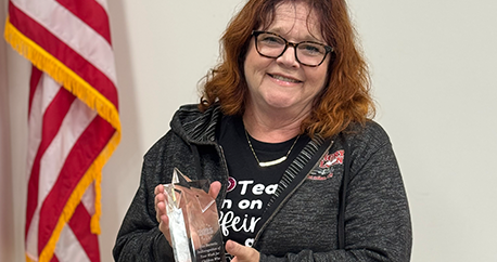 A middle-aged white woman with red hair is standing next to an American flag and holding her award