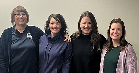 Four white women are standing in front of a white wall and smiling at the camera